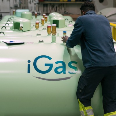 Photo montrant un technicien travaillant sur une citerne de gaz vert clair portant le logo "iGas". Plusieurs citernes similaires sont alignées à l'arrière-plan dans un environnement industriel lumineux, mettant en avant le contrôle et la maintenance des é